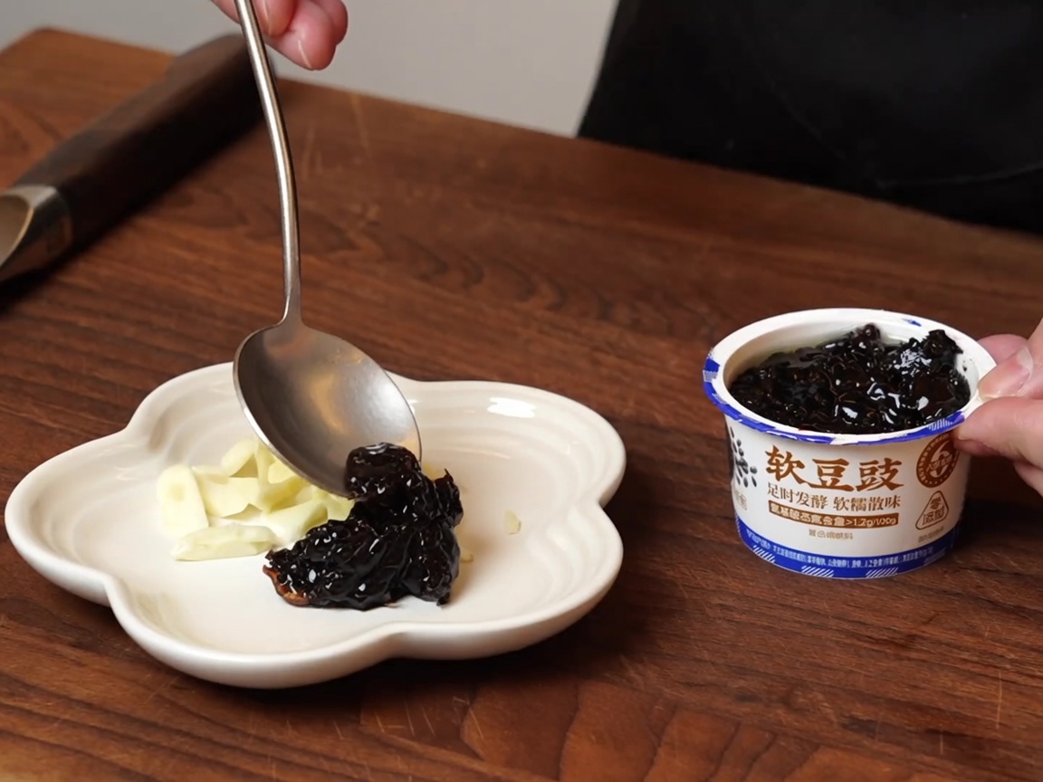 A spoon scooping moist fermented black beans from a container onto a small white plate that holds sliced garlic.