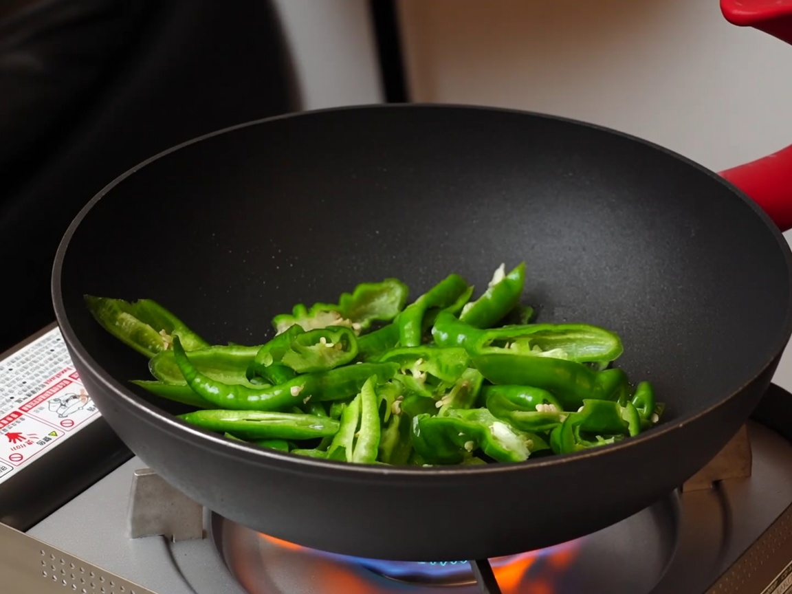 Sliced green peppers being stir-fried in a dark non-stick pan over a portable gas stove.
