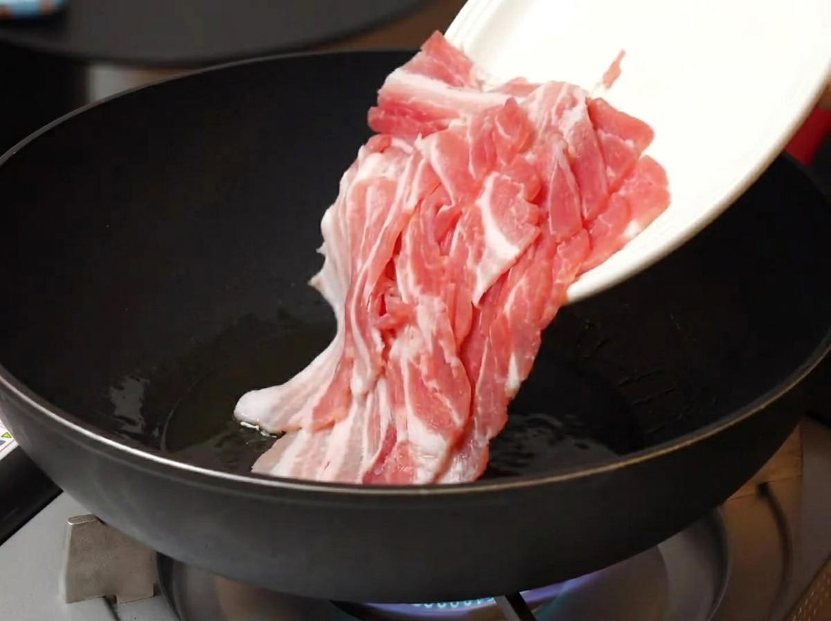 Thinly sliced raw pork belly being added from a white plate into a hot, oiled pan.