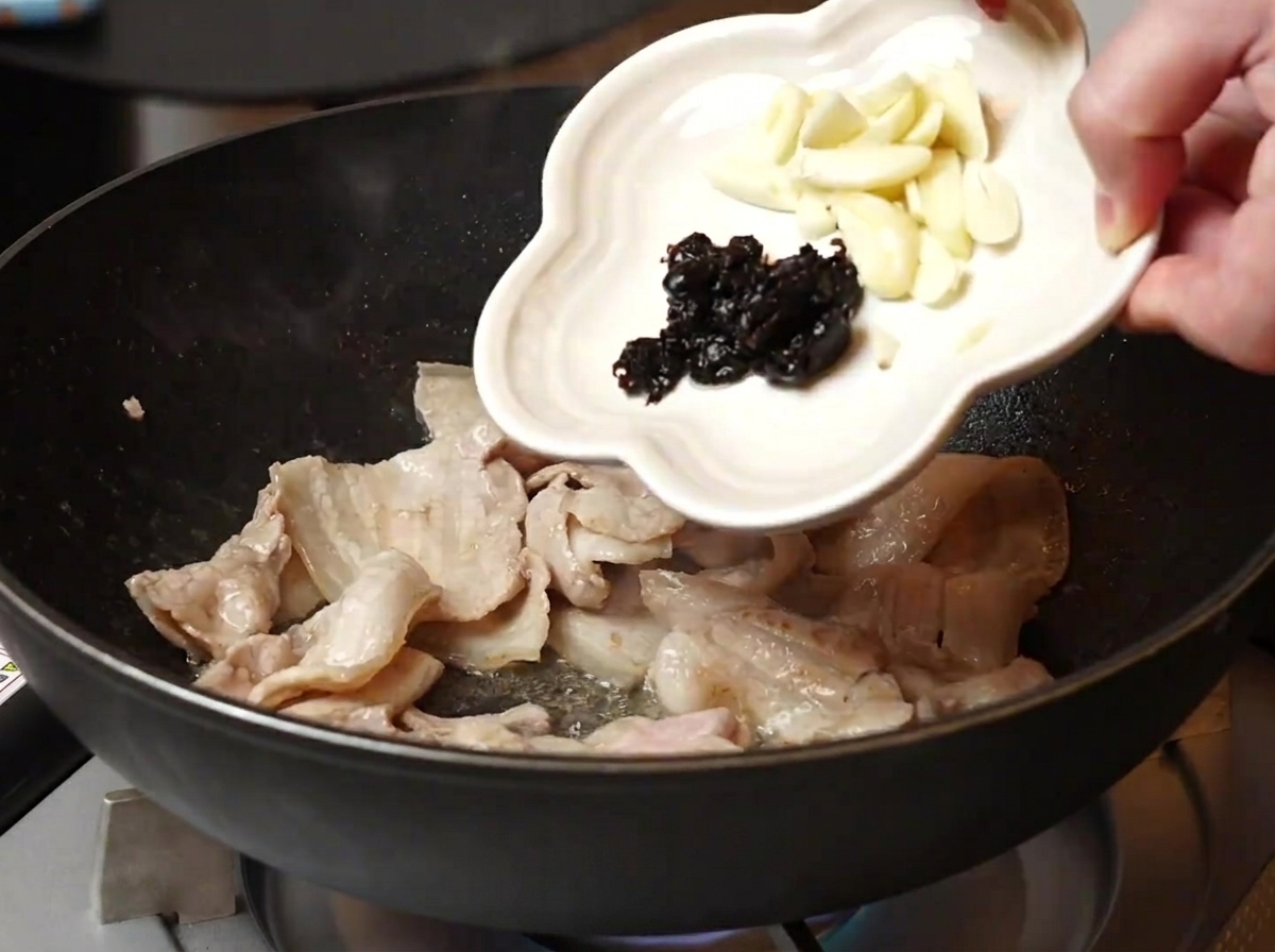 Sliced garlic and fermented black beans being poured from a small white dish into a pan with cooked pork belly.