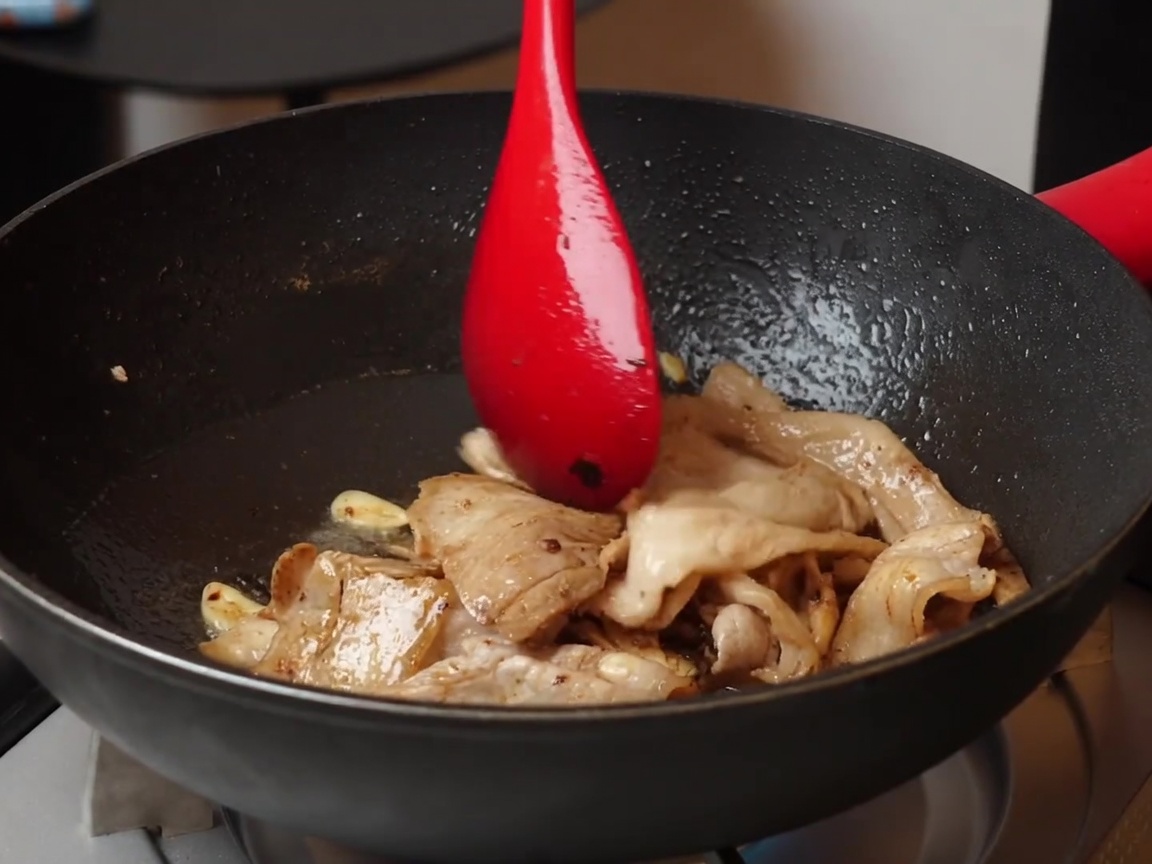 A close-up view of light and dark soy sauce being added to stir-fried pork belly, garlic, and fermented black beans in a non-stick pan.
