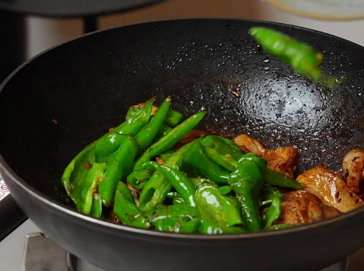 A high-angle shot showing blistered green screw peppers being tossed with the cooked pork belly mixture in a large wok.