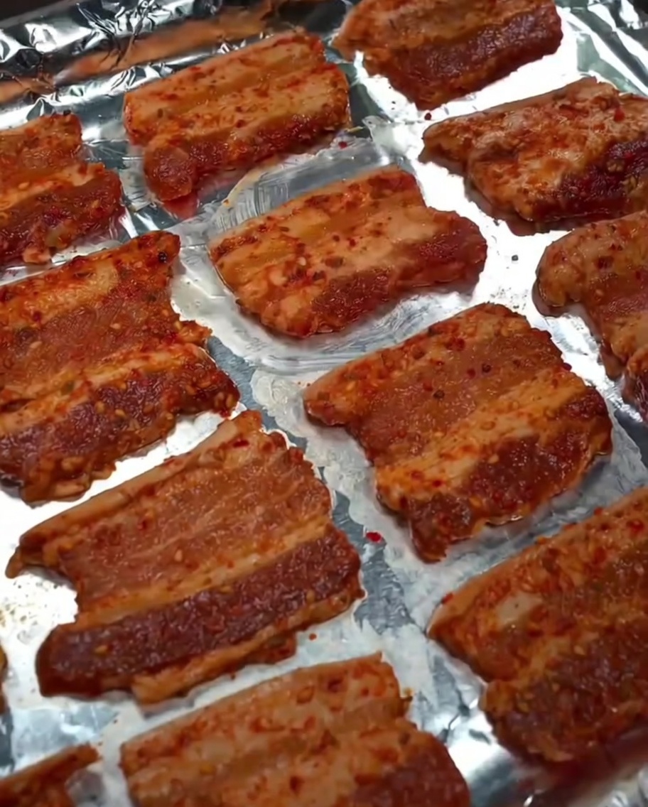 Cooking oil being misted over the raw pork belly slices on the baking sheet.