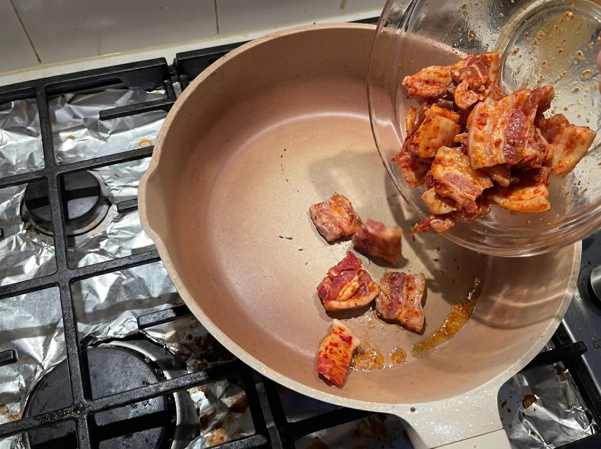 Marinated pork belly slices sliding from a glass bowl into a hot, empty pan to render out their fat.