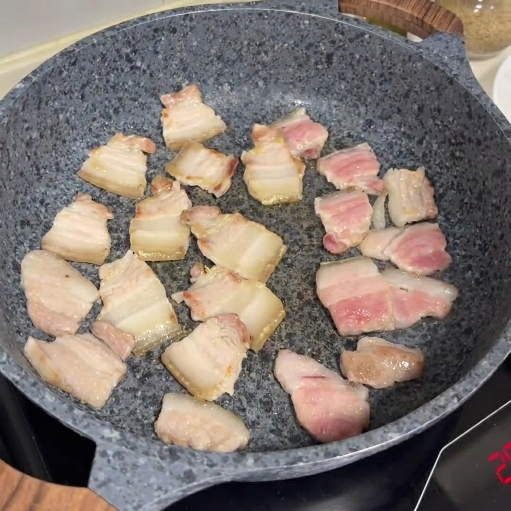 Slices of pork belly frying and browning in a grey stone pan.