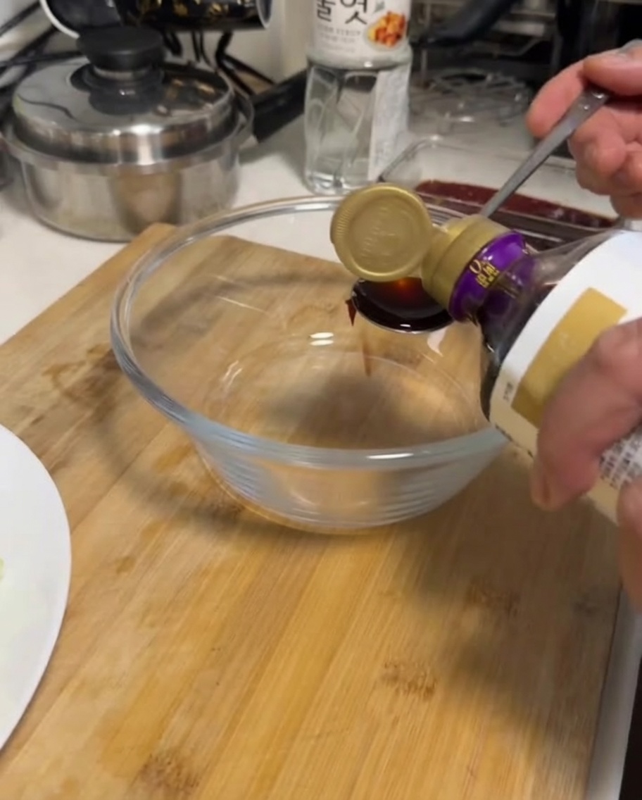 A hand pouring soy sauce from a bottle into a clear glass bowl.