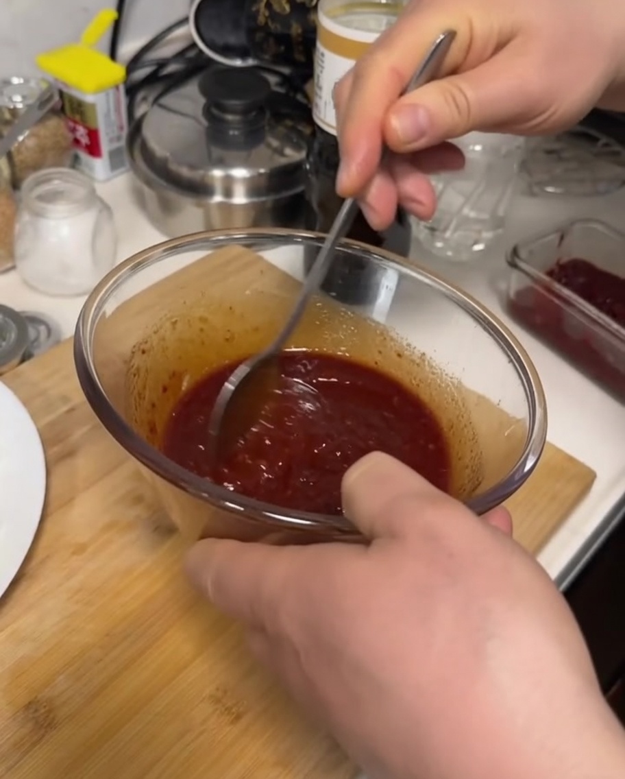 A hand using a metal spoon to mix a dark red, thick sauce inside a clear glass bowl.
