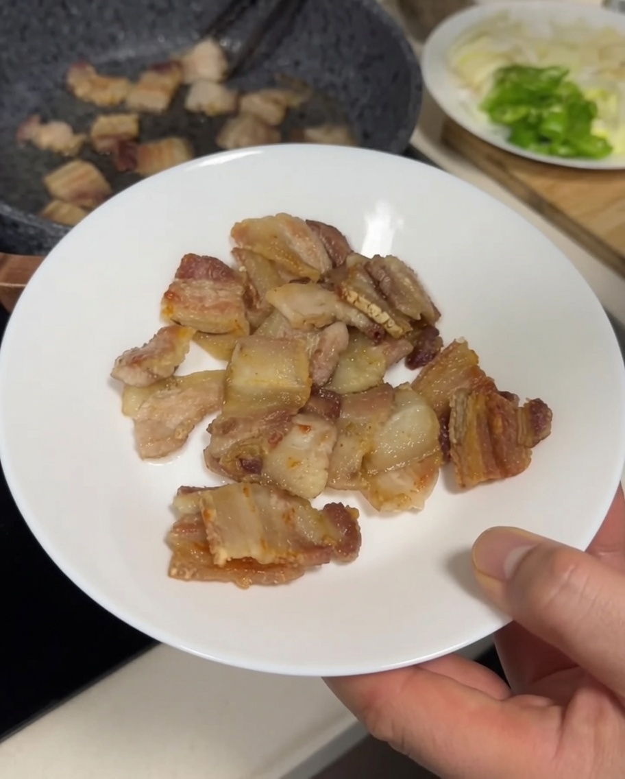 Golden-brown, crispy pieces of pork belly being transferred from a frying pan to a white plate with metal tongs.