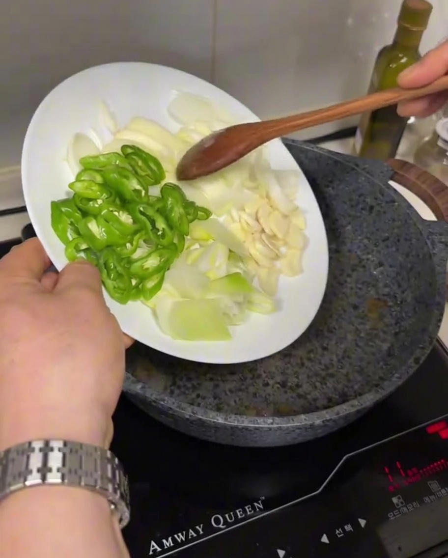 Sliced green chilies, white onions, and garlic being added from a plate into a hot, oiled frying pan.
