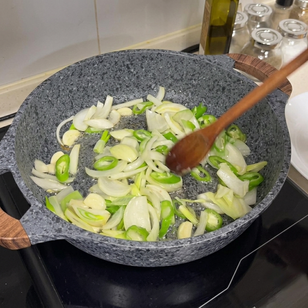 Onions, green chilies, and garlic being stir-fried in a hot pan with a wooden spoon.