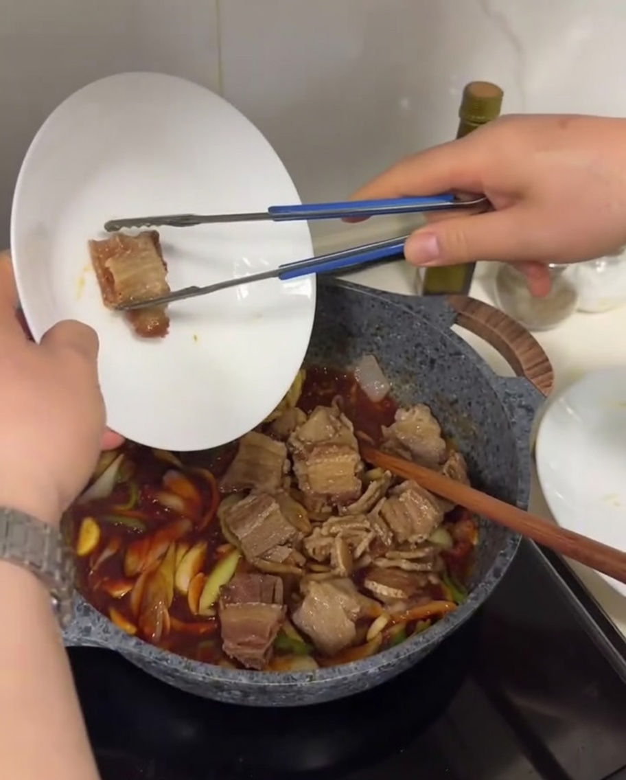 Slices of crispy pan-fried pork belly being added into a pan with sautéed onions, peppers, and a rich, bubbling red chili sauce.