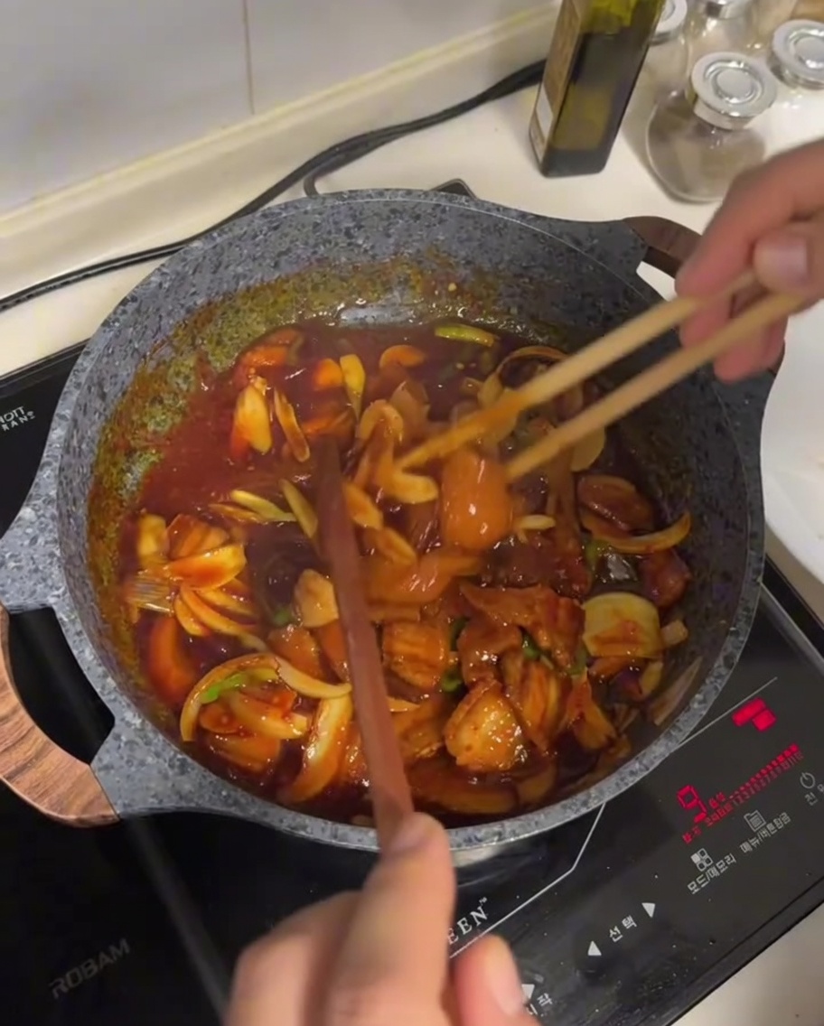 Pork belly and vegetable mixture being stir-fried in a dark, glossy red sauce using long wooden chopsticks in a non-stick pan.