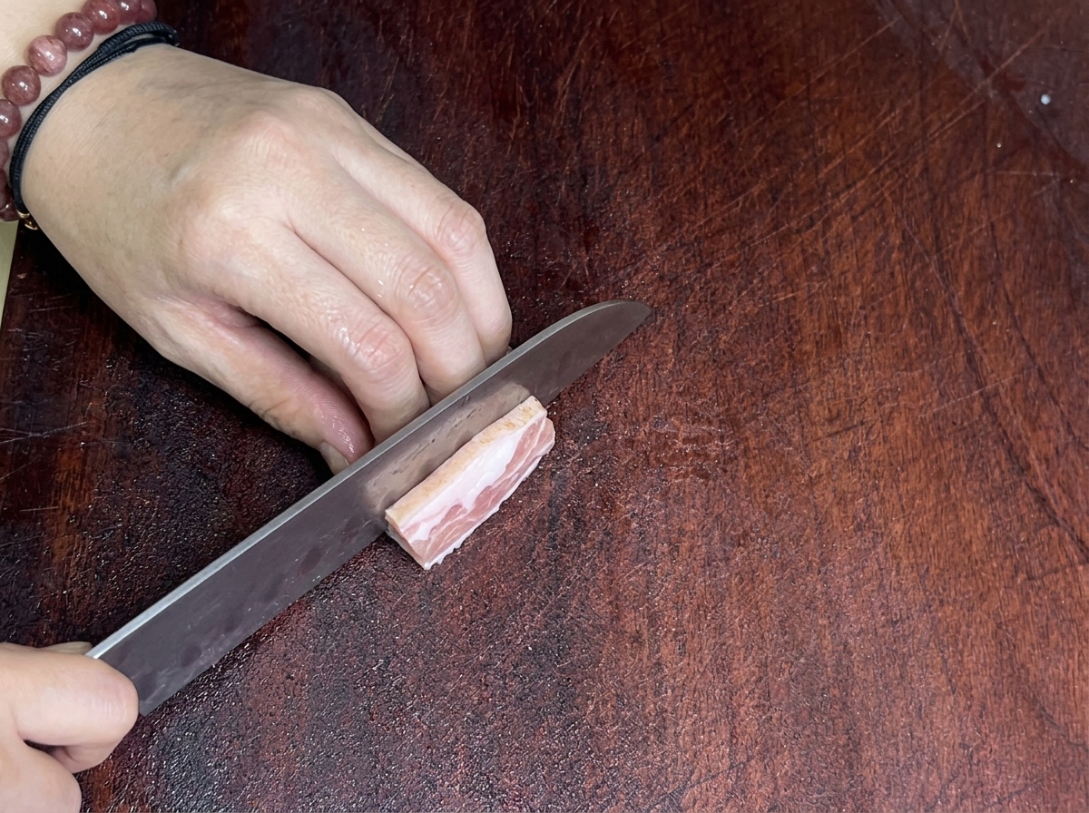 A close-up of a sharp knife slicing through a piece of pork belly on a wooden cutting board.