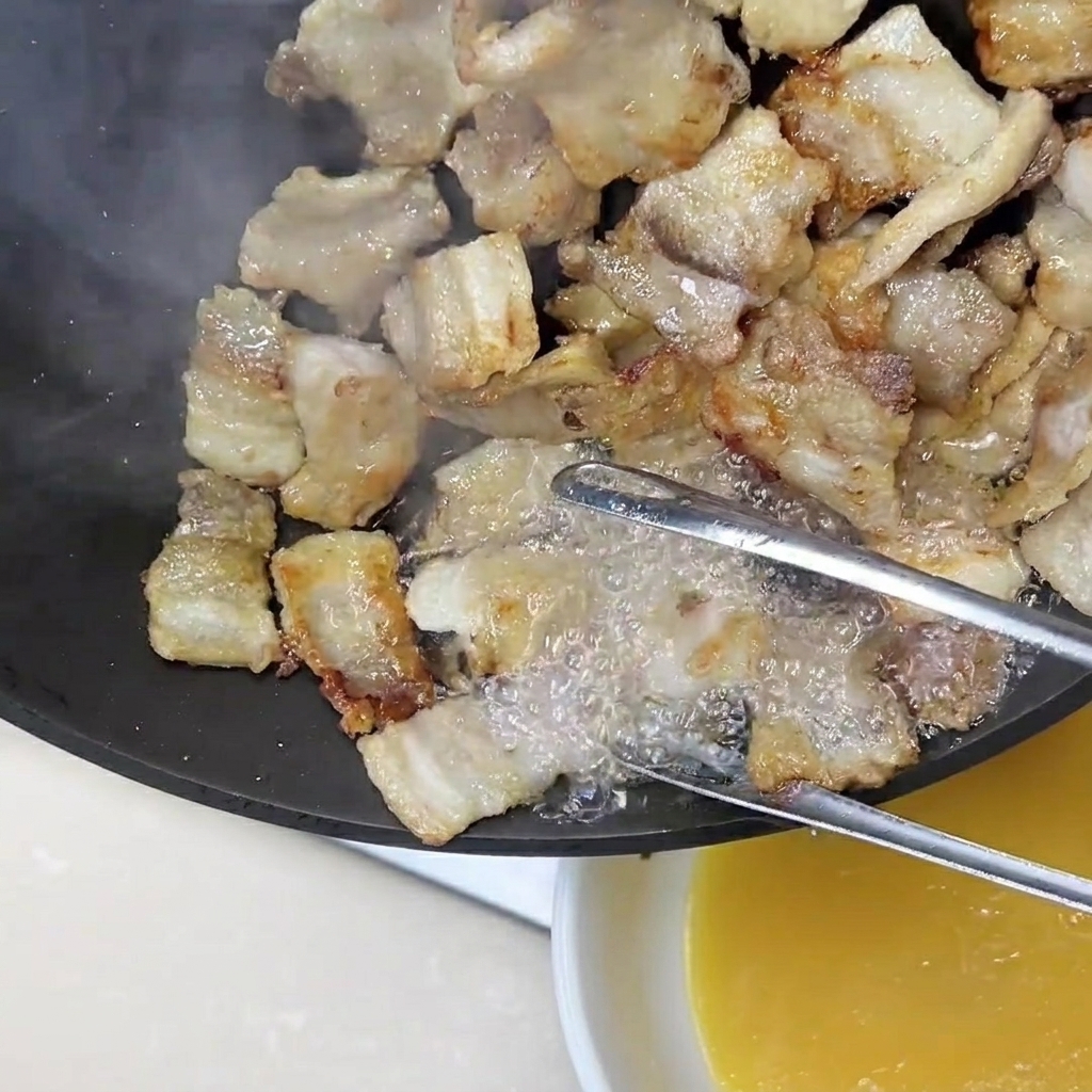 Tongs holding pork belly slices while excess golden fat is poured out of the pan.
