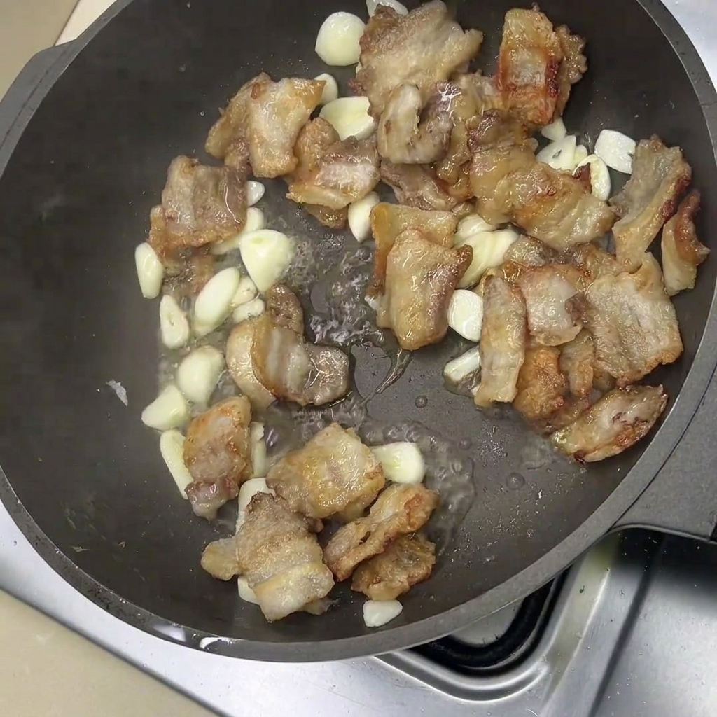 Fried pork belly slices and whole garlic cloves sizzling in a pan.