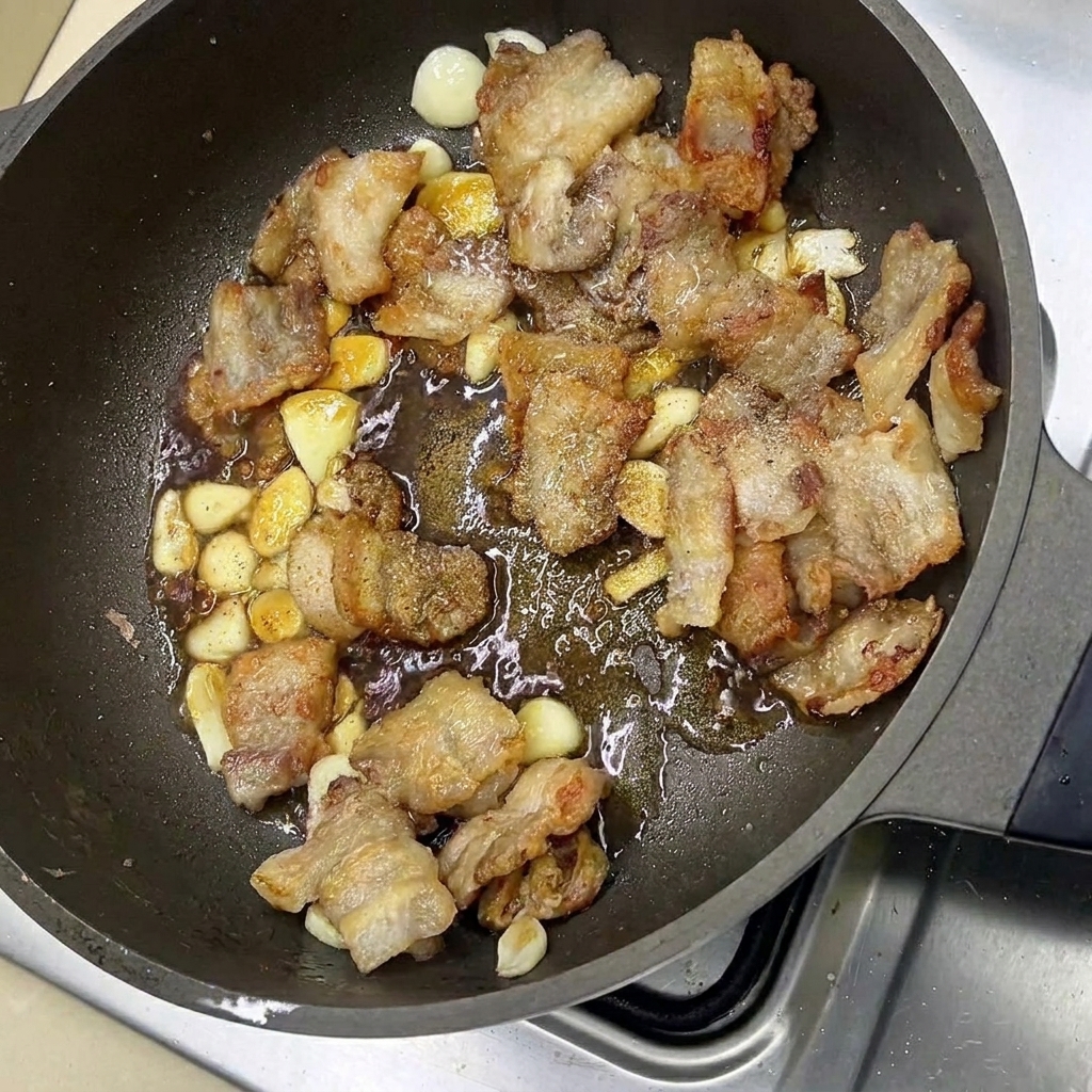 Glazed pork belly and garlic cloves being tossed in a frying pan with savory seasonings.