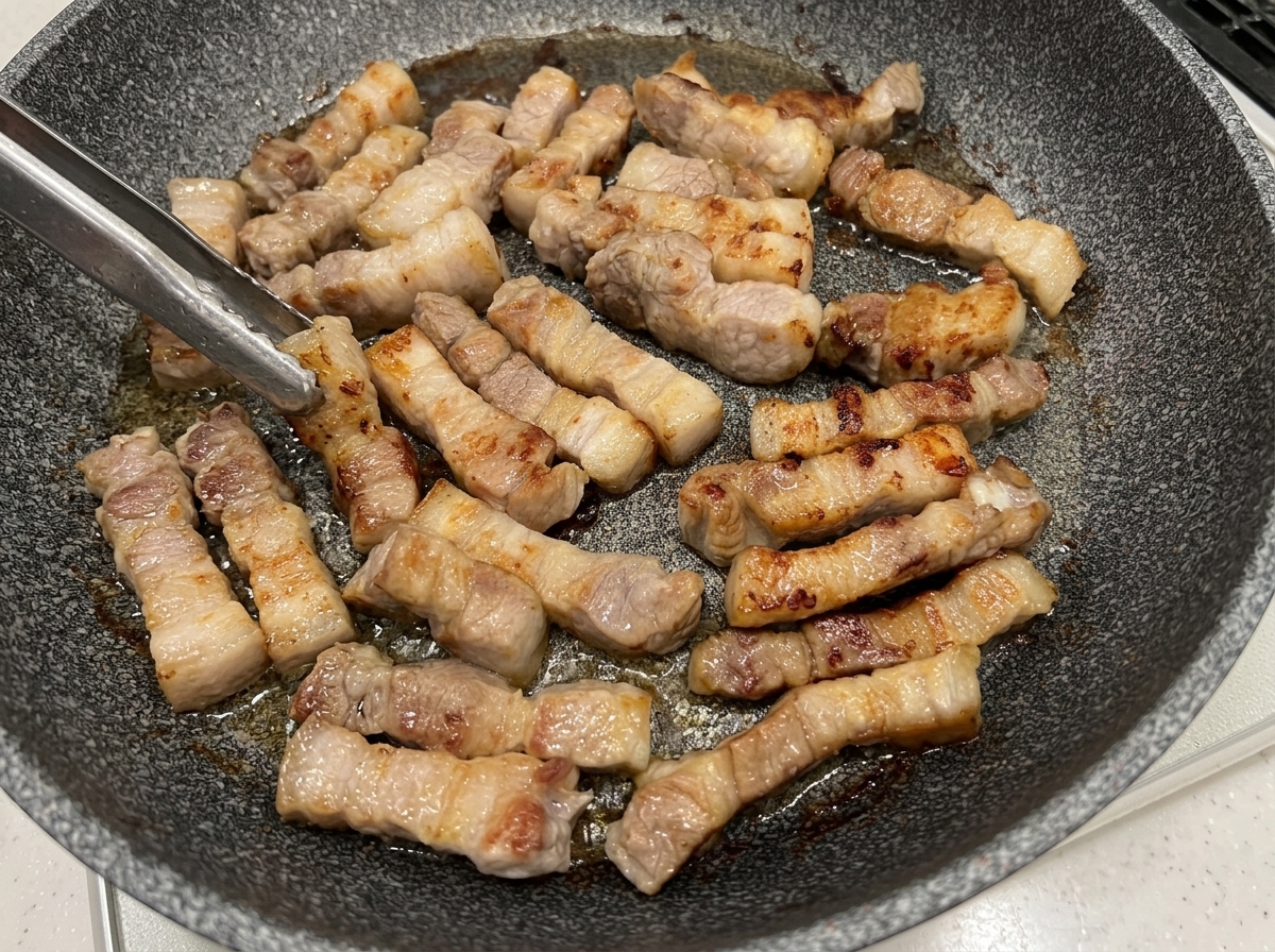 Pork belly pieces being turned with tongs in a pan, showing golden-brown rendered surfaces.