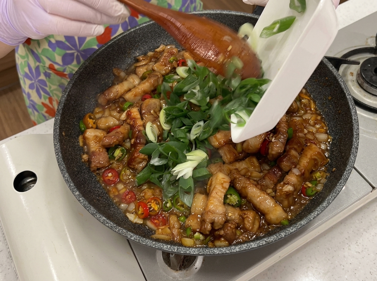 Green chilies, red chilies, and scallions being poured from a small dish into the pan of stir-frying pork belly.