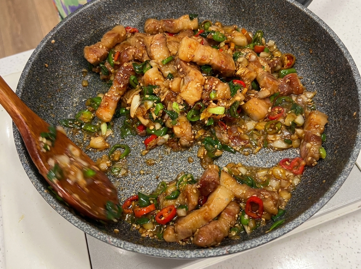 Pork belly pieces, chili, and scallions being mixed in the pan with two wooden spoons.
