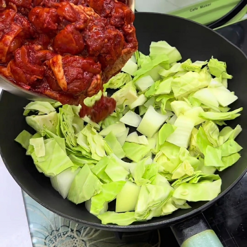 Marinated chicken pieces layered over cabbage and onions in a frying pan.