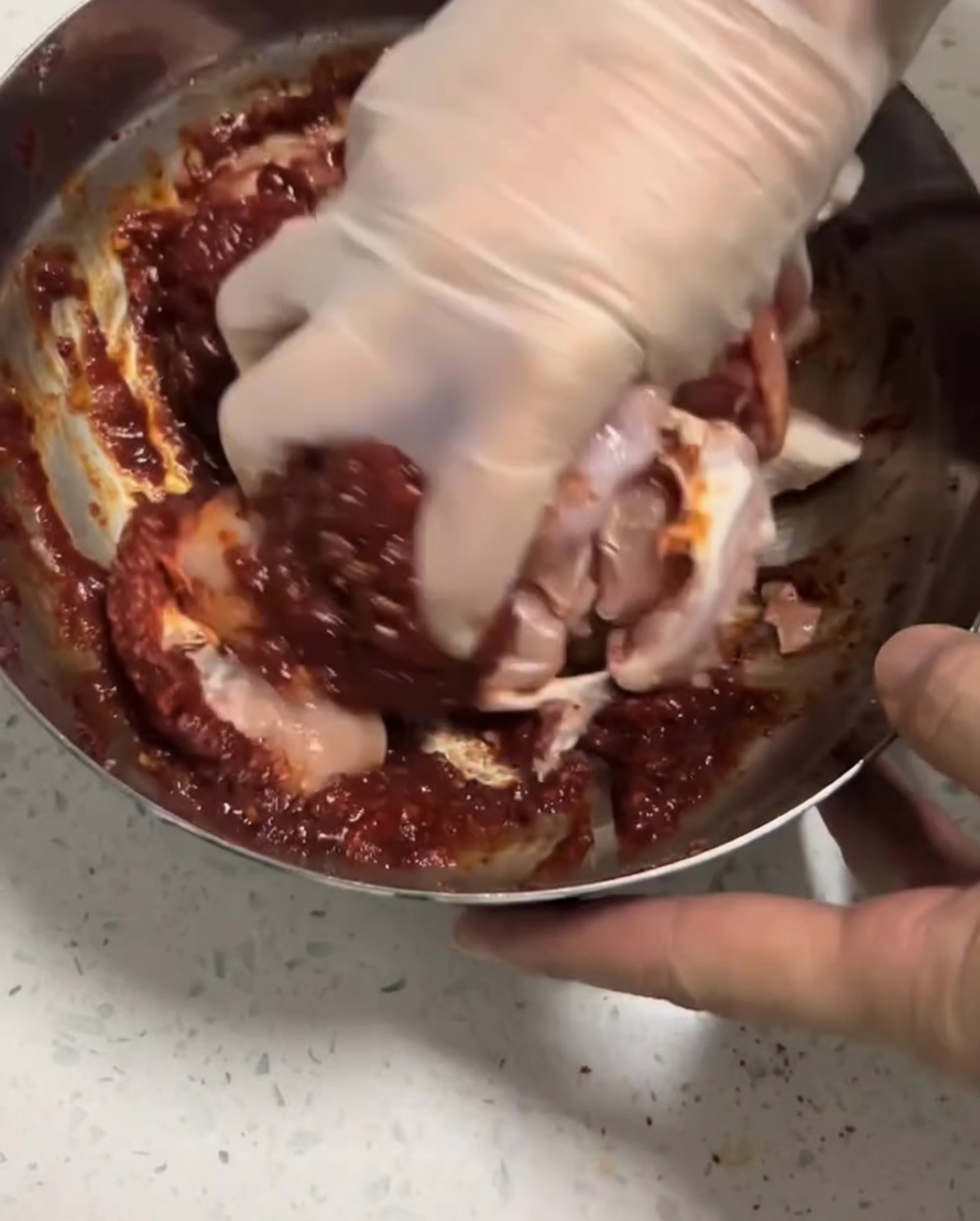 Raw chicken pieces being massaged by a gloved hand into the red marinade inside a bowl.
