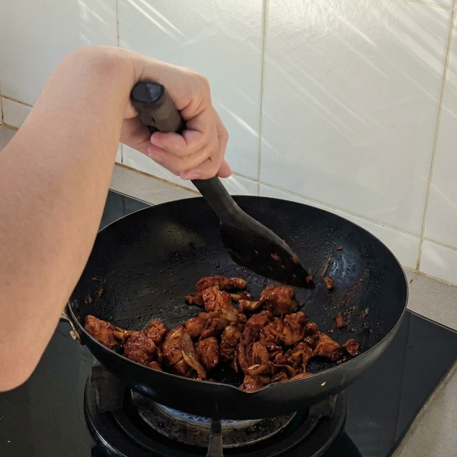 Chicken and onion pieces being stir-fried in a black wok with a spatula, coated in a dark spicy sauce.