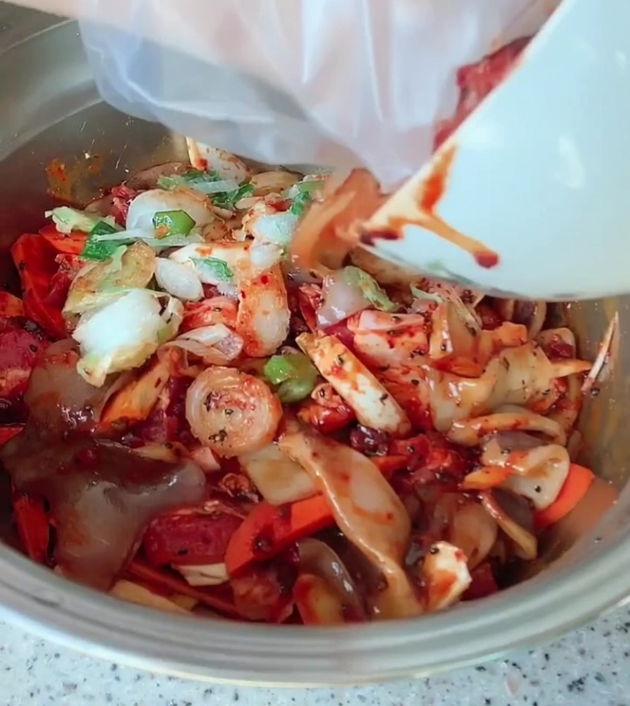 A large metal bowl containing sliced pork belly, colorful vegetables, and green onions being coated in a vibrant red spicy sauce.
