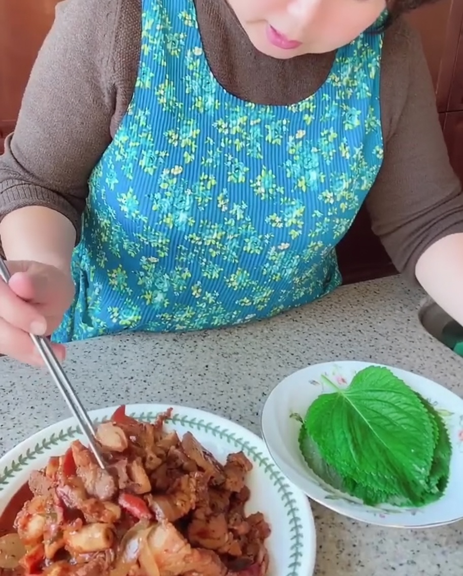 A person using chopsticks to place a piece of cooked pork belly and squid onto a fresh green perilla leaf.