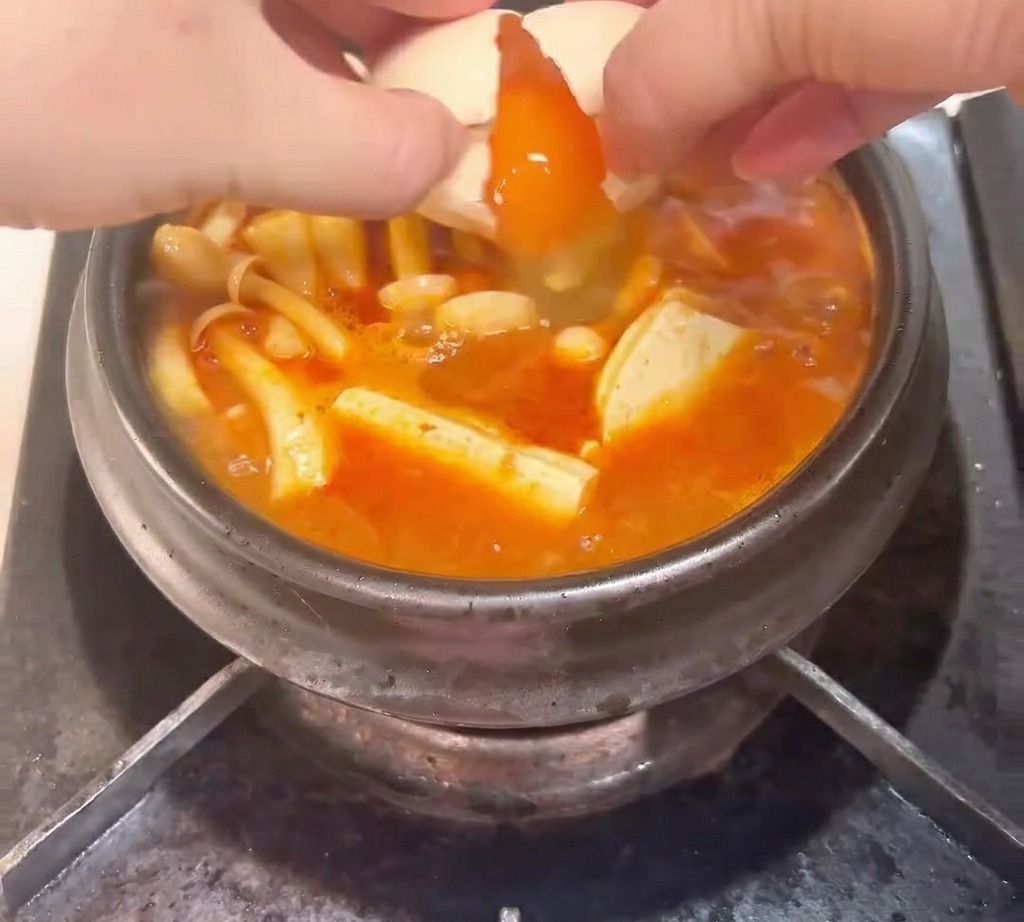 A raw egg being cracked into a bubbling clay pot filled with kimchi, tuna, mushrooms, and tofu.