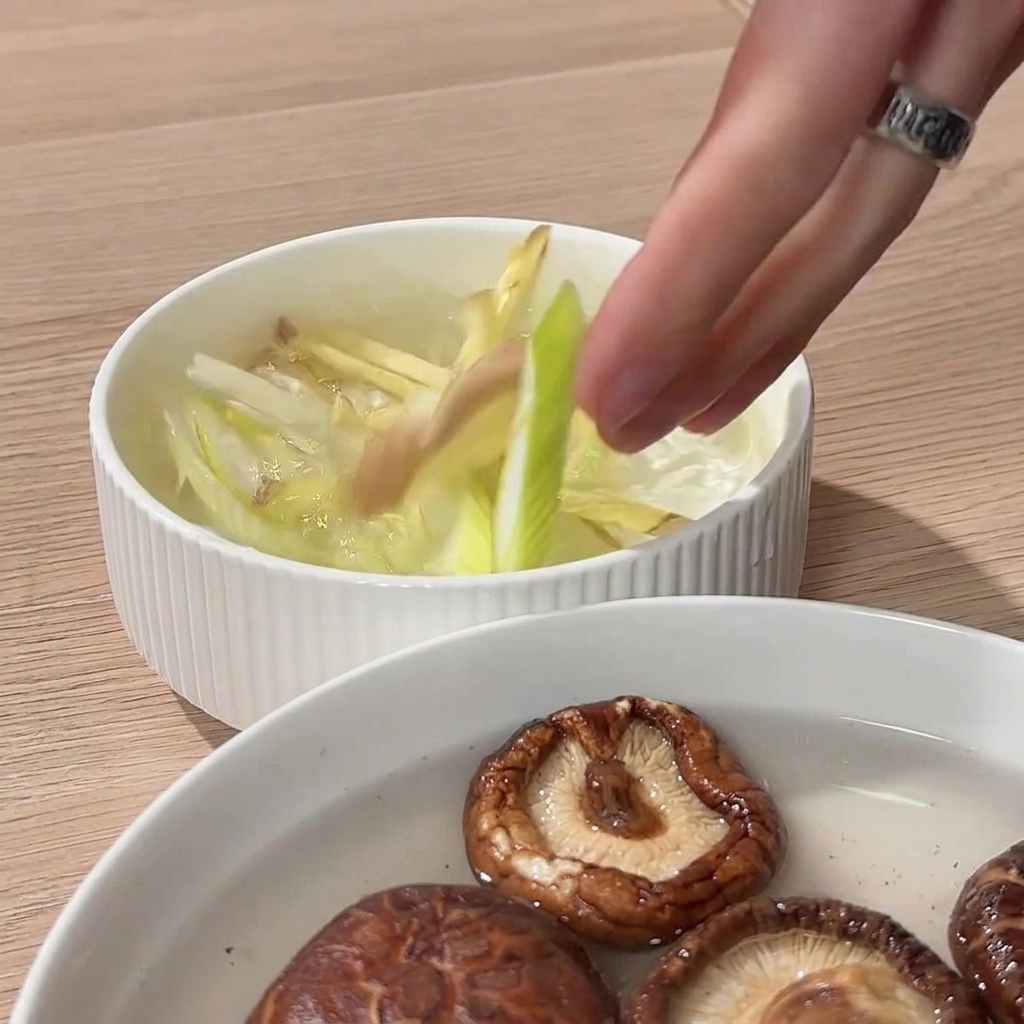 A hand squeezing sliced ginger and green onions in a bowl of water to create a flavorful aromatic infusion.