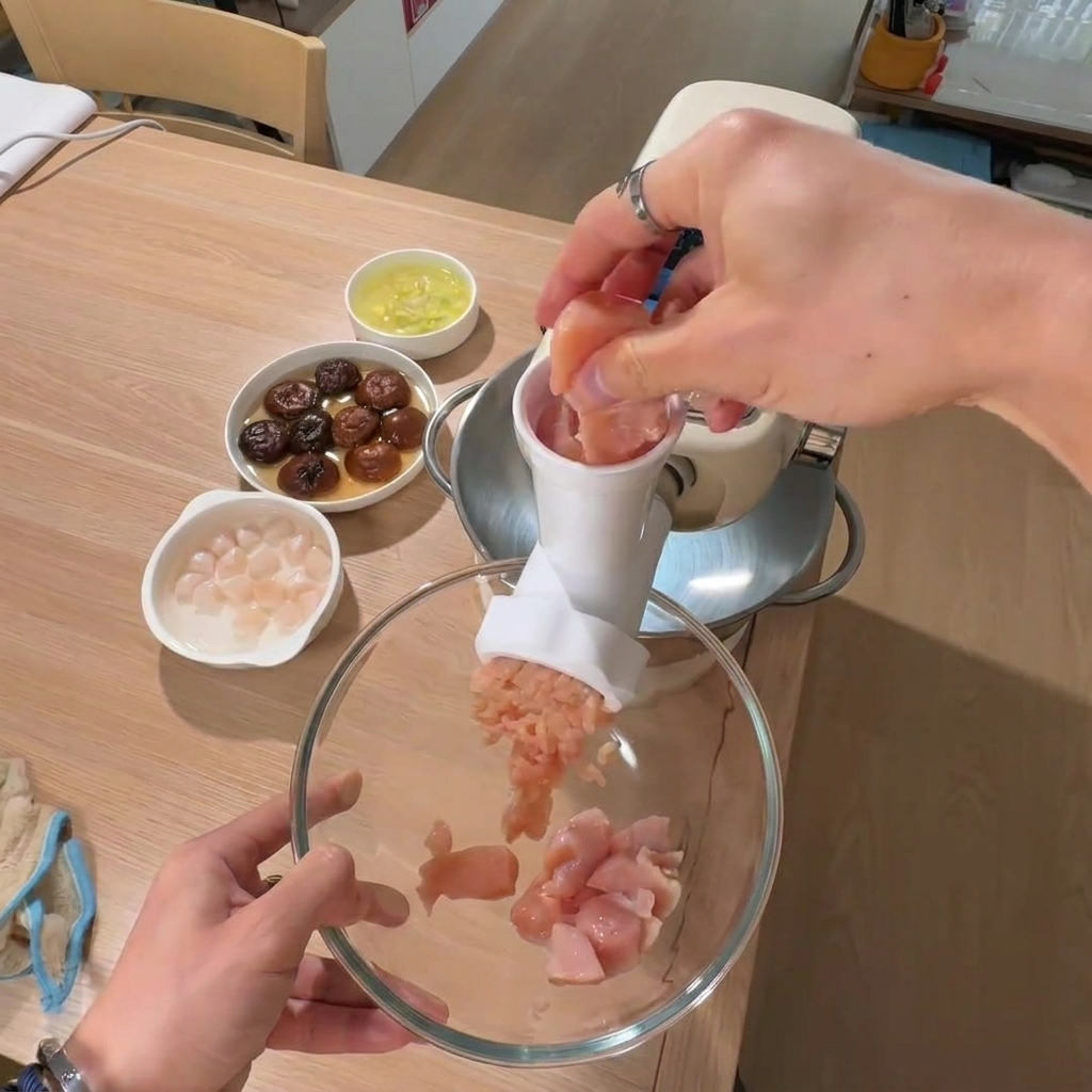A hand placing pieces of chicken breast into a large bowl with bowls of soaked mushrooms and scallops in the background.