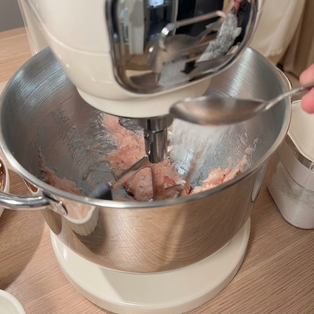 A spoonful of seasoning being added to the minced chicken in the stand mixer bowl, with other ingredients visible on the table.
