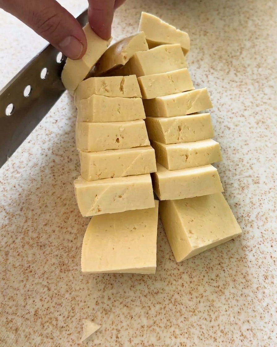 A block of tofu being cut into neat, uniform pieces on a board.