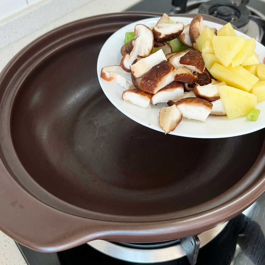 Chopped potatoes, zucchini, and shiitake mushrooms being added to a brown earthenware pot.