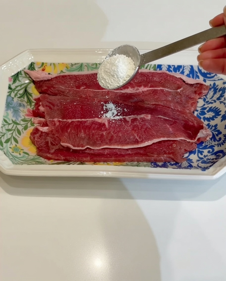 A spoonful of starch being added to thin, raw beef slices on a patterned plate.