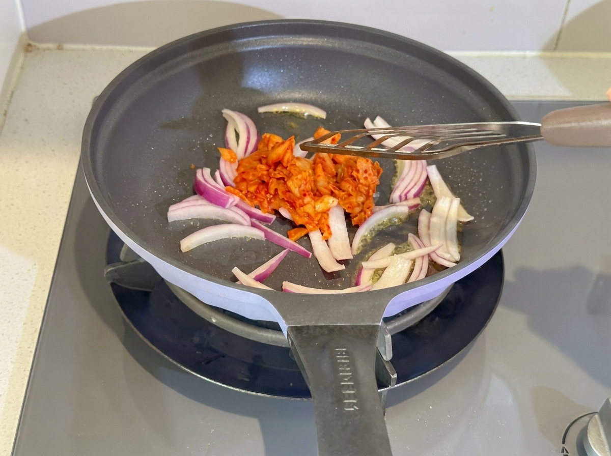 Kimchi and red sauce being stirred into the sautéed onions in a pan.