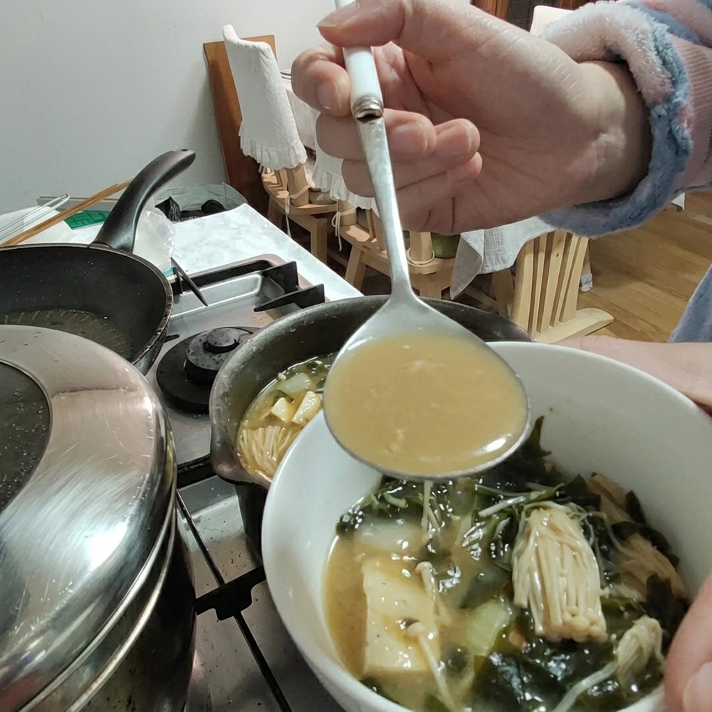 Soybean paste being added to the stew using a ladle, with tofu and enoki mushrooms visible in the bowl.