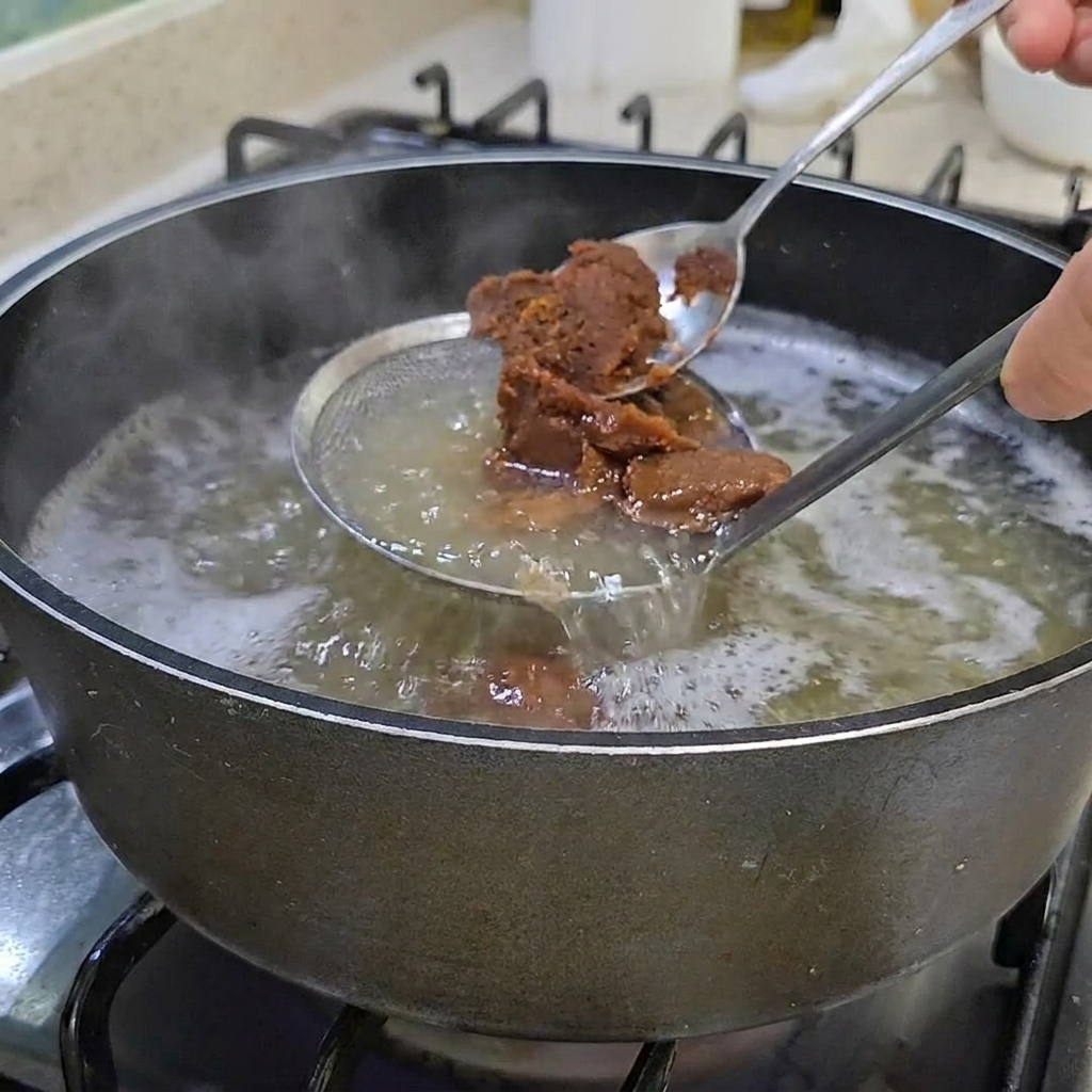 Two spoonfuls of thick, brown Korean soybean paste being stirred into a pot of clear broth.