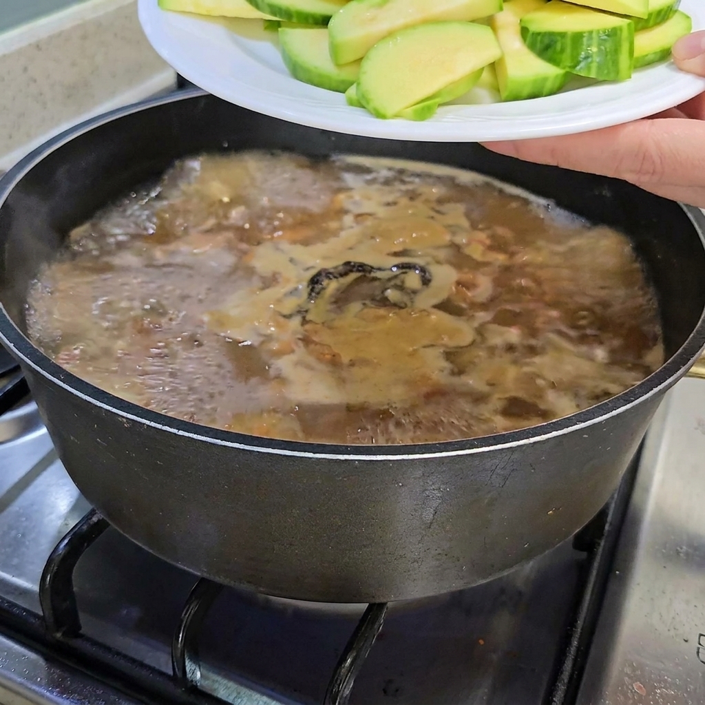 Sliced zucchini and a bunch of enoki mushrooms being added to the simmering stew pot.