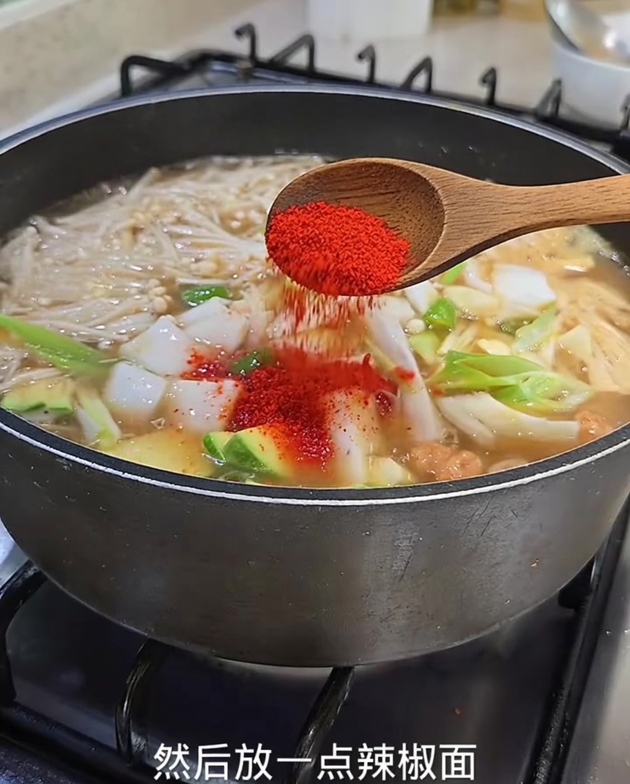 A close-up view of vibrant red chili powder being sprinkled from a wooden spoon into the simmering stew containing zucchini, onions, and enoki mushrooms.