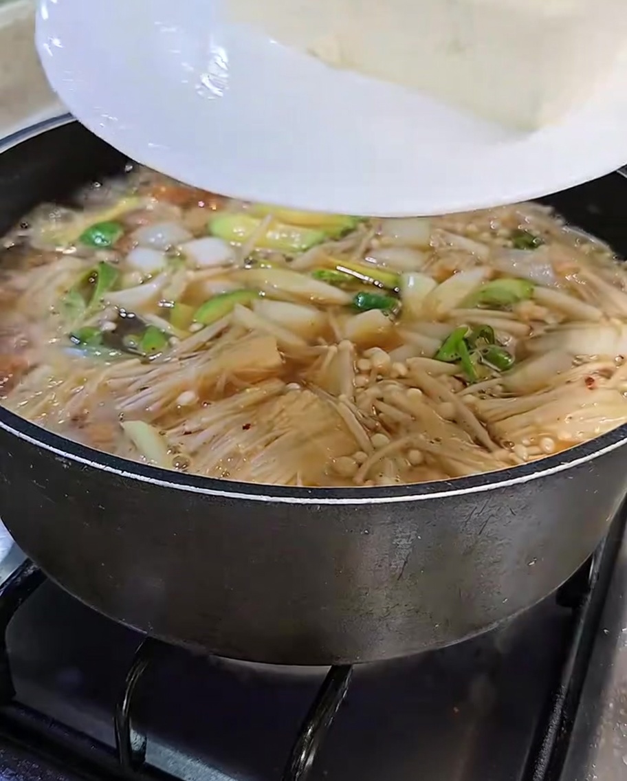 Slices of white tofu being carefully transferred from a white plate into the bubbling, flavorful soybean paste stew.