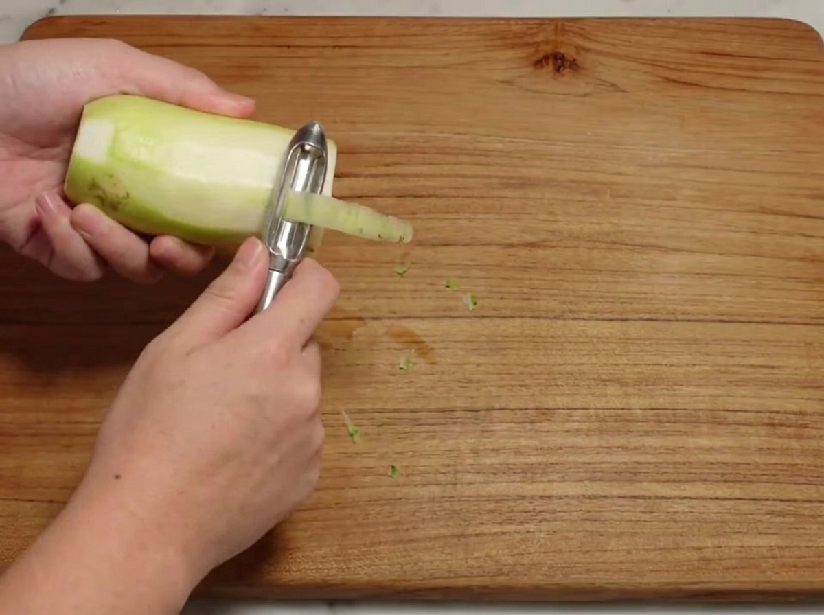 A raw white radish being peeled on a wooden cutting board.