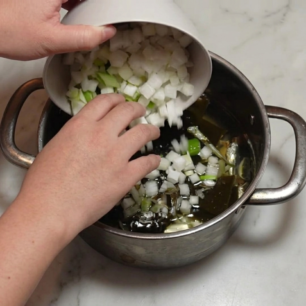 Chopped radish and green onion being poured from a bowl into a pot with anchovies and kombu.