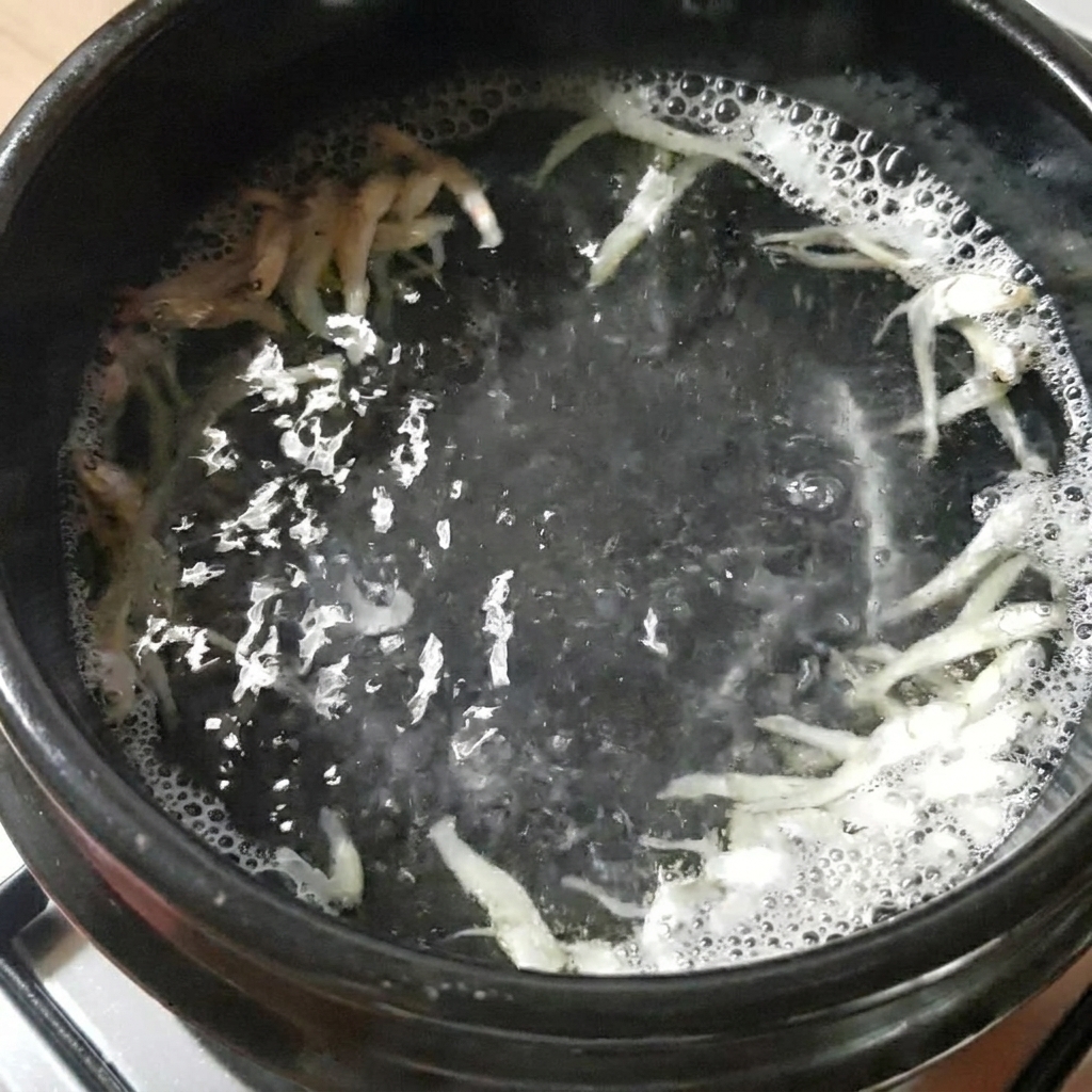 A black stone pot containing water, dried anchovies, and kelp, boiling on a stovetop.