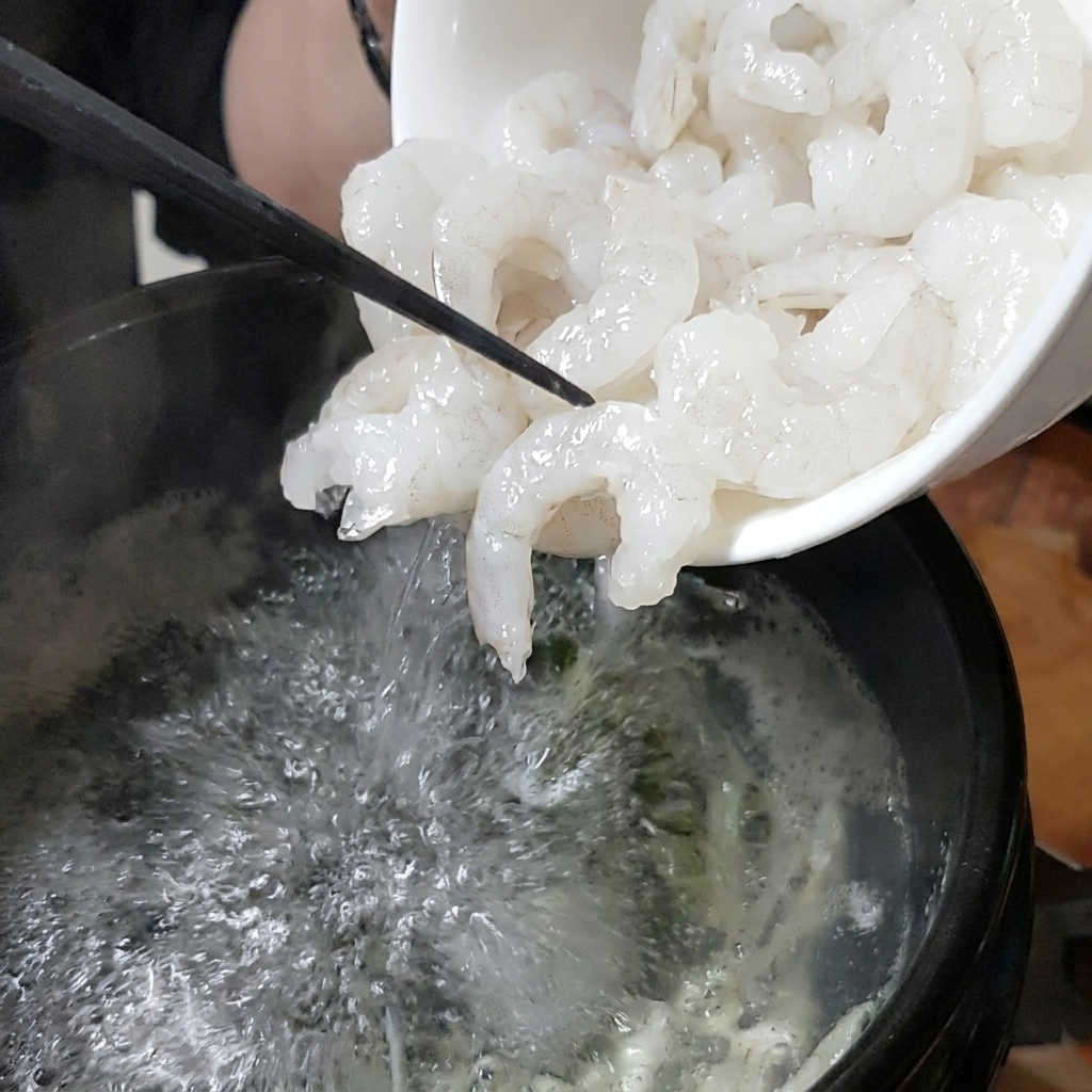 A white bowl pouring fresh shrimp into a bubbling stone pot of broth.
