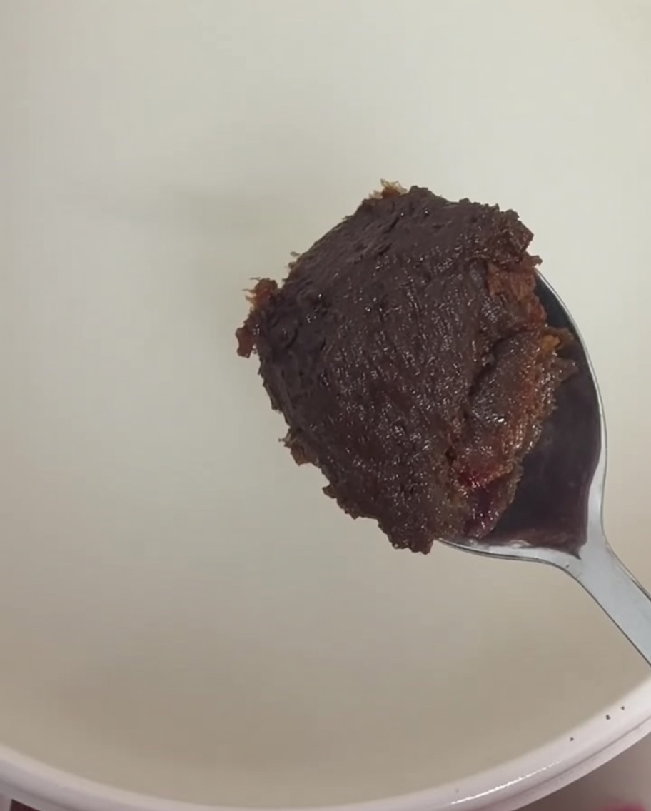 A spoonful of thick brown soybean paste and dark red chili paste being stirred into the rice water broth.