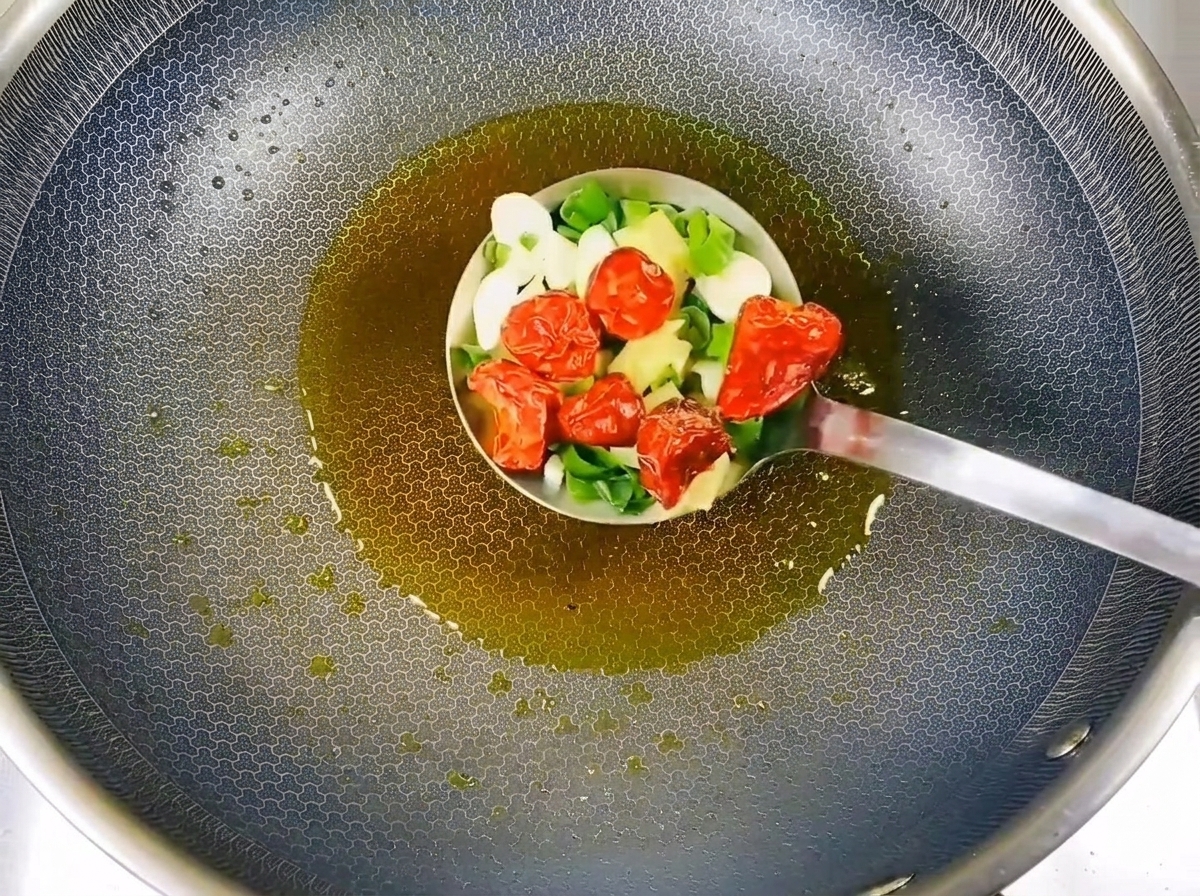 Chopped green onions, garlic slices, and dried red chilies being stir-fried in hot oil inside a metal ladle.