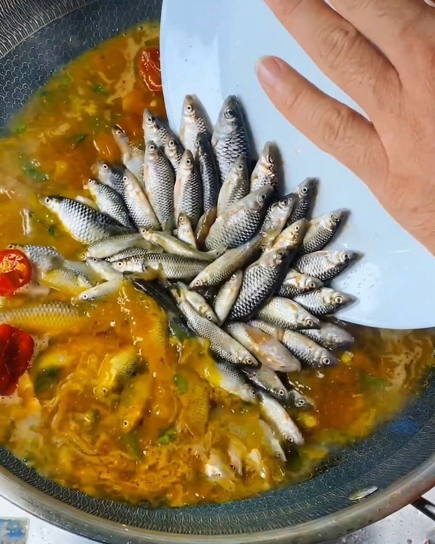 A hand pouring liquid seasoning from a bottle over the arranged fish in the wok.