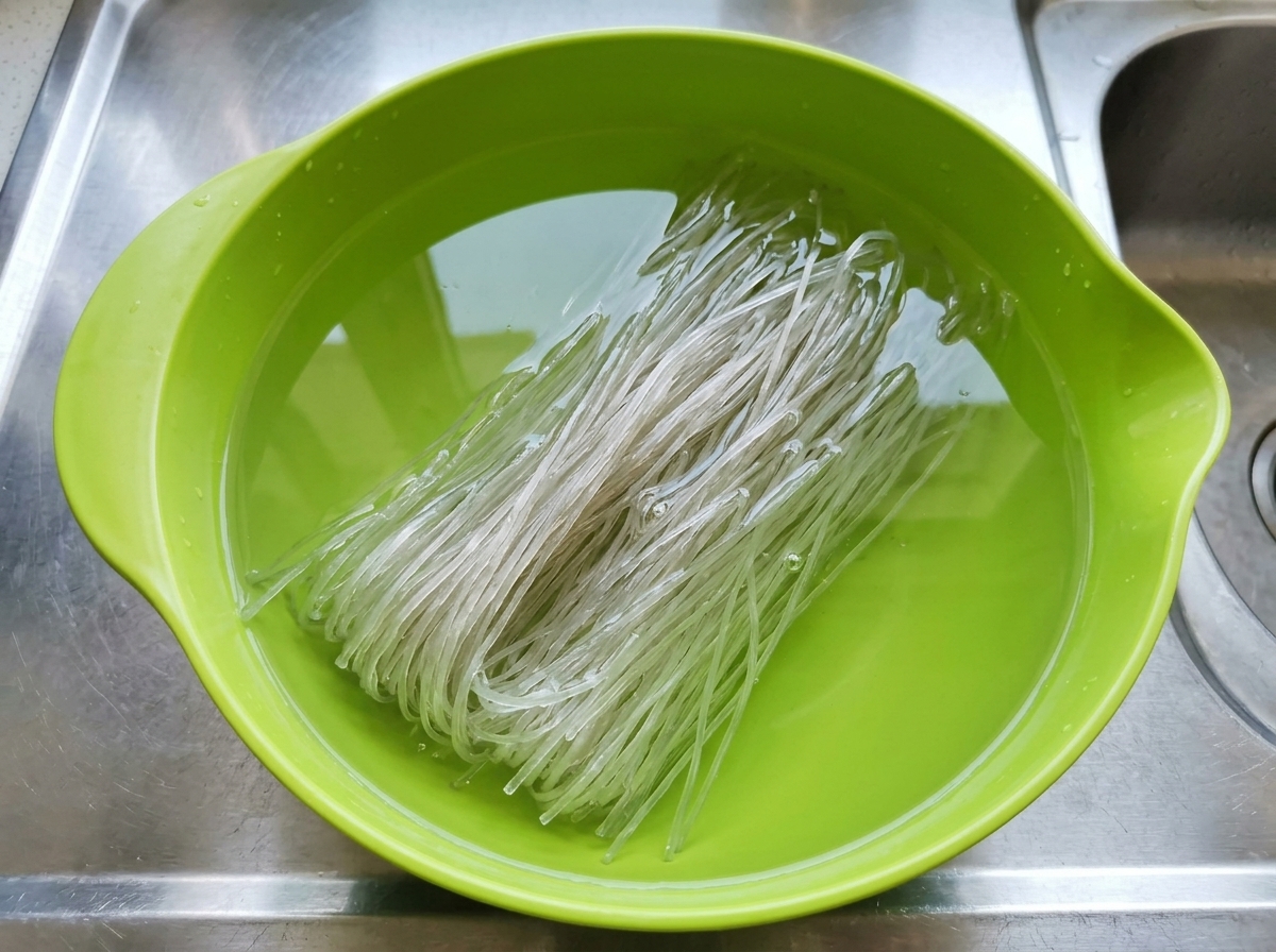 Dry glass noodles completely submerged in cold water inside a bright green mixing bowl.