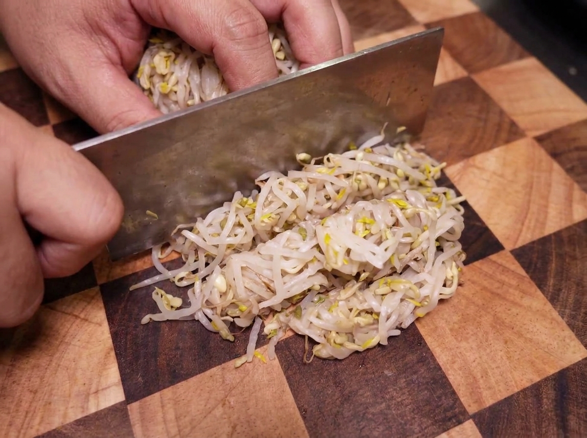 A hand using a large metal cleaver to chop a compressed bundle of mung bean sprouts on a checkered wooden board.